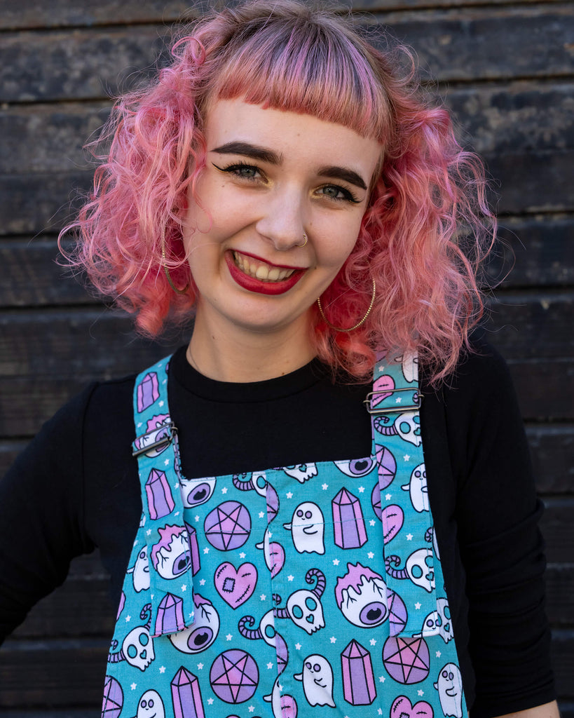 A woman with pink curly hair and fringe smiles warmly at the camera. She is wearing a turquoise pinafore dress covered in a vibrant "Pretty Spooky" print featuring ghosts, skulls, crystals, and hearts. The dress is by Run & Fly, a small ethical slow fashion brand. The black backdrop contrasts with the colourful outfit, drawing attention to the fun and quirky design.