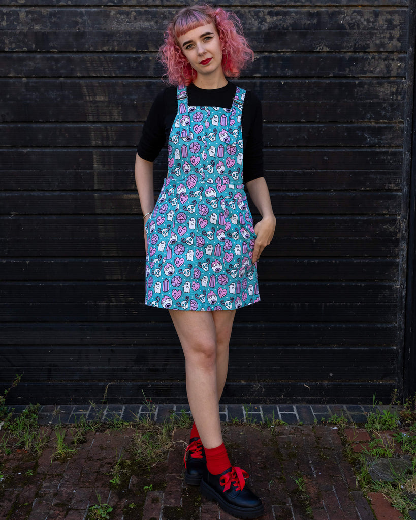 A woman with pink curly hair stands confidently in a turquoise "Pretty Spooky" pinafore dress by Run & Fly. The dress is covered in a pastel print featuring ghosts, skulls, hearts, and crystals. She pairs it with black platform shoes and red socks. Run & Fly is a small ethical slow fashion company known for its playful and unique designs.