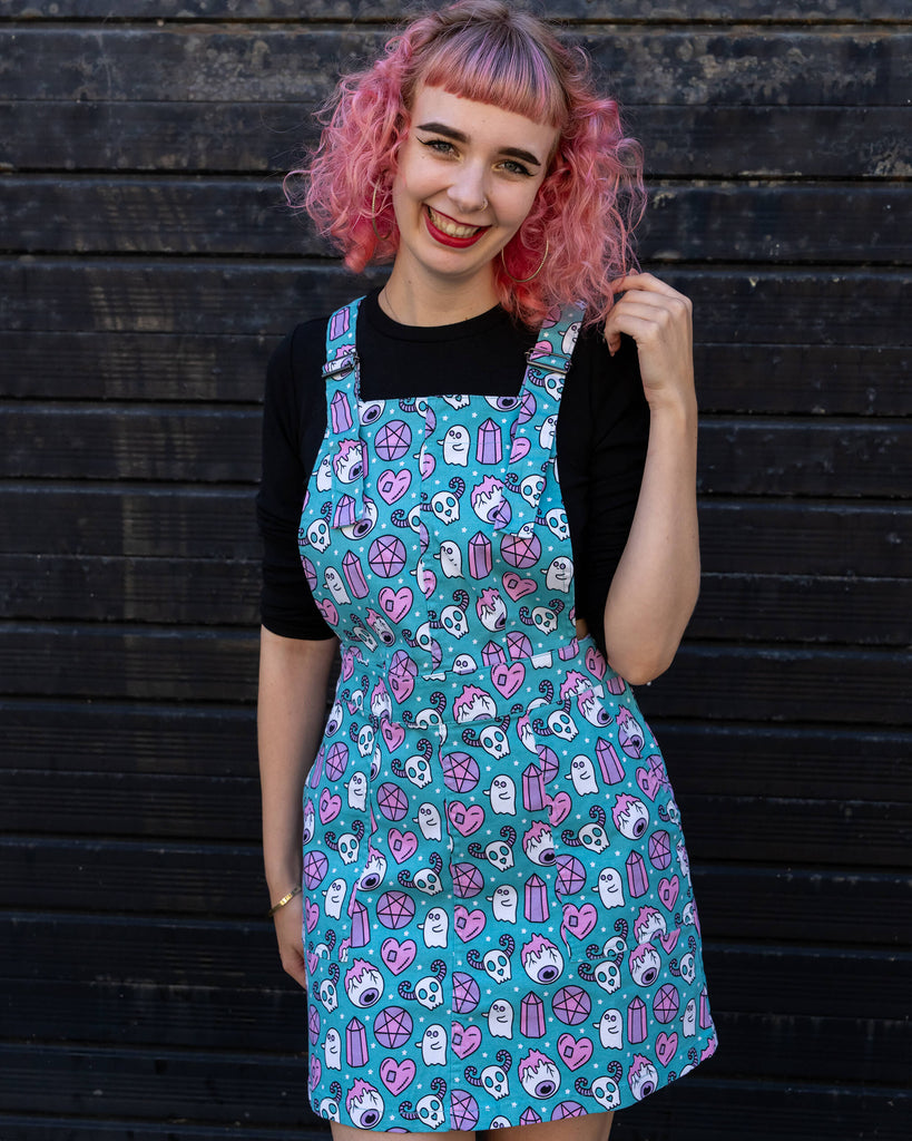 A smiling woman with pink curly hair poses in a vibrant "Pretty Spooky" pinafore dress by Run & Fly. The turquoise dress features a playful print of pastel skulls, ghosts, and crystals, paired with a black long-sleeve top. Run & Fly is a small ethical slow fashion company creating fun, alternative fashion.