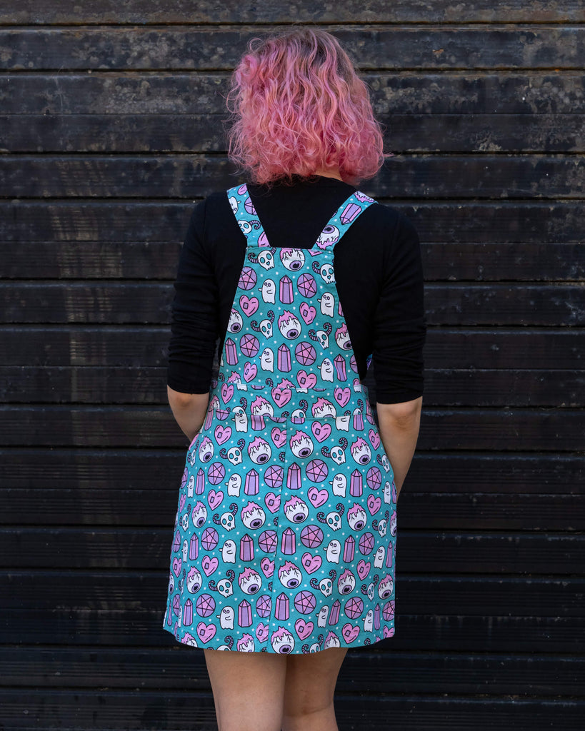 Back view of a woman with pink curly hair wearing a "Pretty Spooky" turquoise pinafore dress by Run & Fly. The dress features an all-over print of pastel ghosts, skulls, crystals, and hearts. The playful design stands out against the black long-sleeve top she is wearing. Run & Fly is a small ethical slow fashion company.