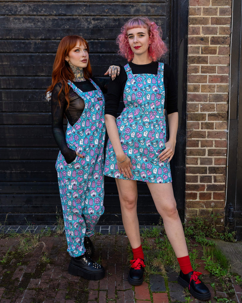 Two women pose in front of a brick and black wooden wall wearing coordinating "Pretty Spooky" outfits by Run & Fly. The pink-haired model wears a turquoise pinafore dress, and the red-haired model wears matching dungarees. Both outfits feature a quirky print of ghosts, skulls, and crystals. Run & Fly is a small ethical slow fashion brand.