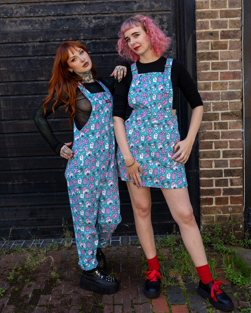 Two women stand together wearing matching "Pretty Spooky" outfits by Run & Fly. One model, with pink curly hair, wears a turquoise pinafore dress, while the other, with long red hair and tattoos, wears turquoise dungarees with the same ghost, crystal, and skull print. Both outfits are styled with black tops. Run & Fly is a small ethical slow fashion company known for its bold, playful prints.