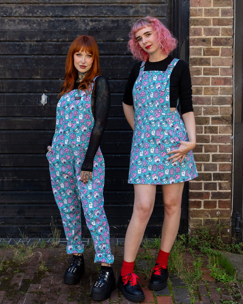 Two women pose together wearing matching outfits from Run & Fly. One wears turquoise dungarees, and the other a matching pinafore dress, both featuring the same quirky, spooky-themed print in pastel pinks and purples. They stand against a brick wall, with one woman sporting pink hair and the other red, adding a pop of colour to the scene.