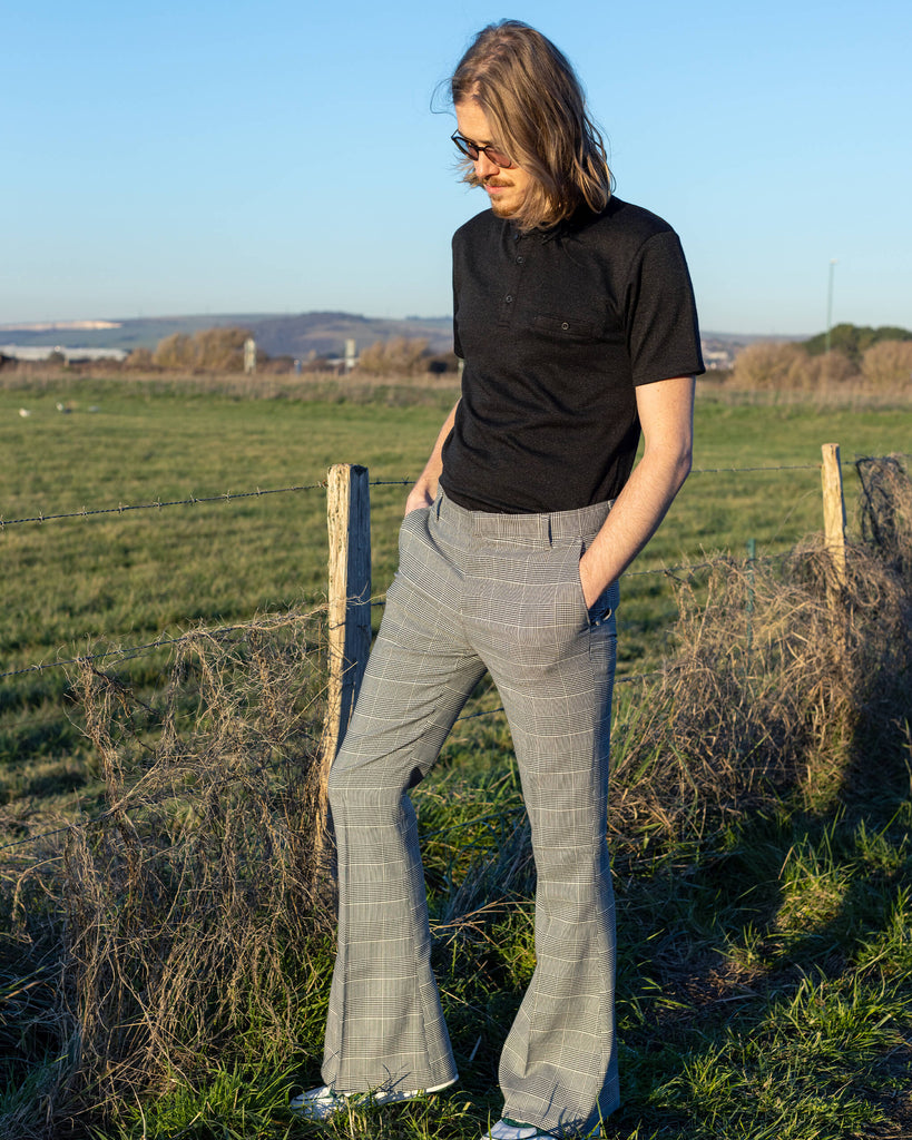 Jack is stood outside near a grass field wearing the Presley prince of Wales check bell bottom trousers with a black short sleeve shirt and white trainers. They have mid length blonde hair with circle polarised glasses. Jack is posing with his hands in the pockets of the trousers and has one leg bent.
