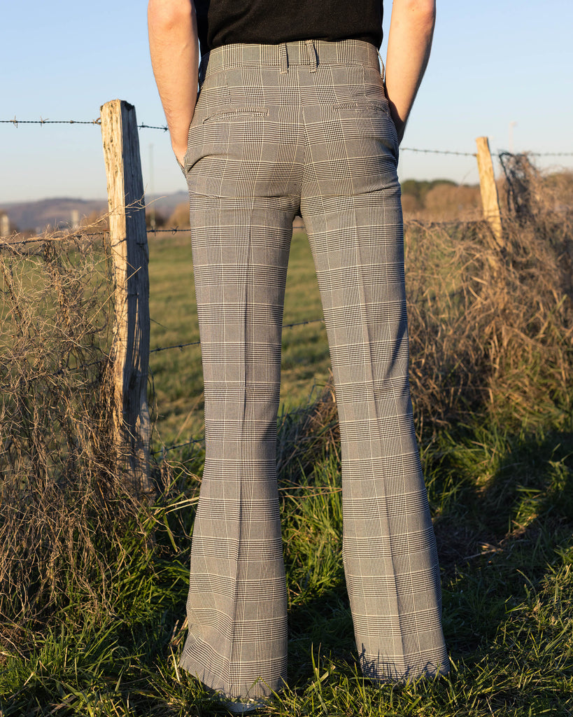 Jack is stood outside near a grass field wearing the Presley prince of Wales check bell bottom trousers with a black short sleeve shirt and white trainers. 
 Jack is stood with his back to the camera and the photo is cropped at the waist