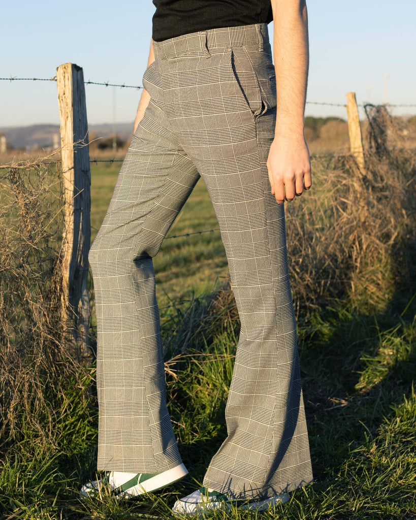 Jack is stood outside near a grass field wearing the Presley prince of Wales check bell bottom trousers with a black short sleeve shirt and white trainers. Jack is posing with his hands in the pockets of the trousers and has one leg bent. The photo is cropped at the waist