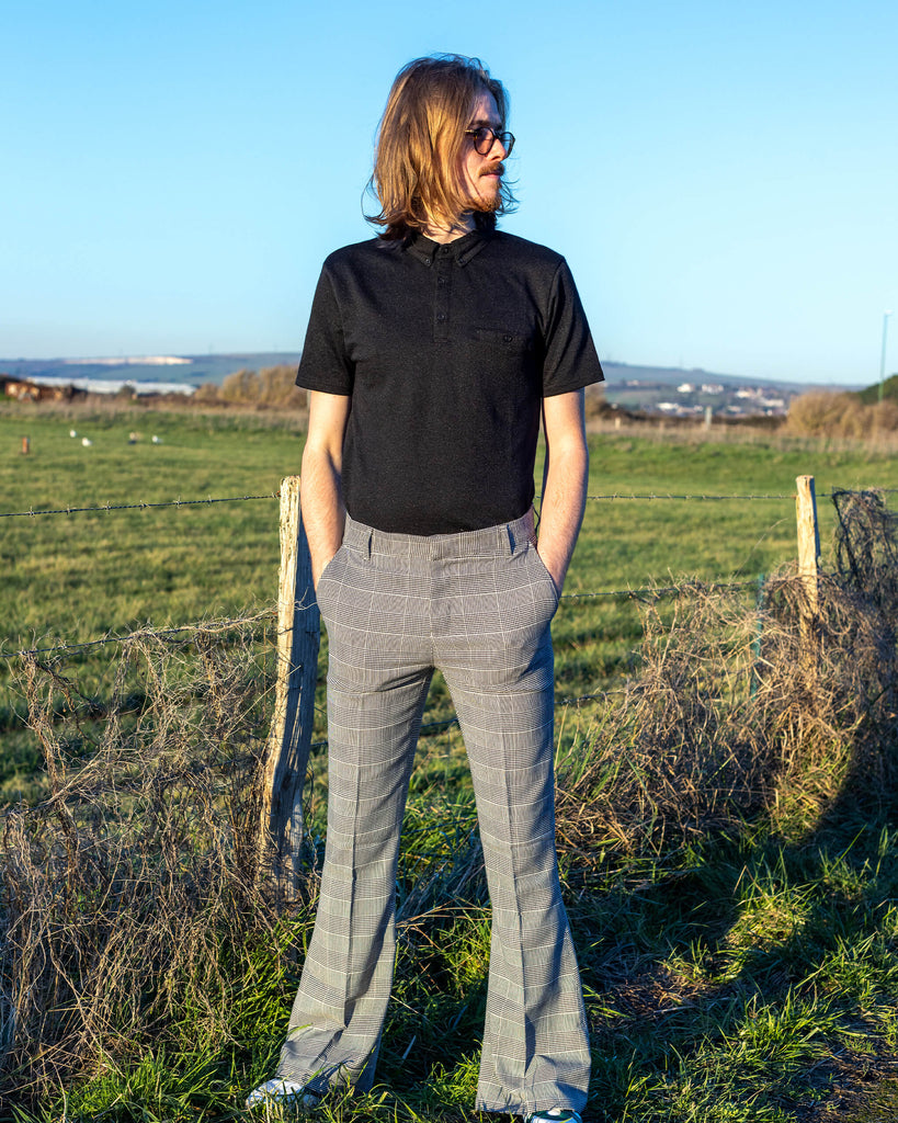 Jack is stood outside near a grass field wearing the Presley prince of Wales check bell bottom trousers with a black short sleeve shirt and white trainers. They have mid length blonde hair with circle polarised glasses. Jack is posing with his hands in the pockets of the trousers and has one leg bent.