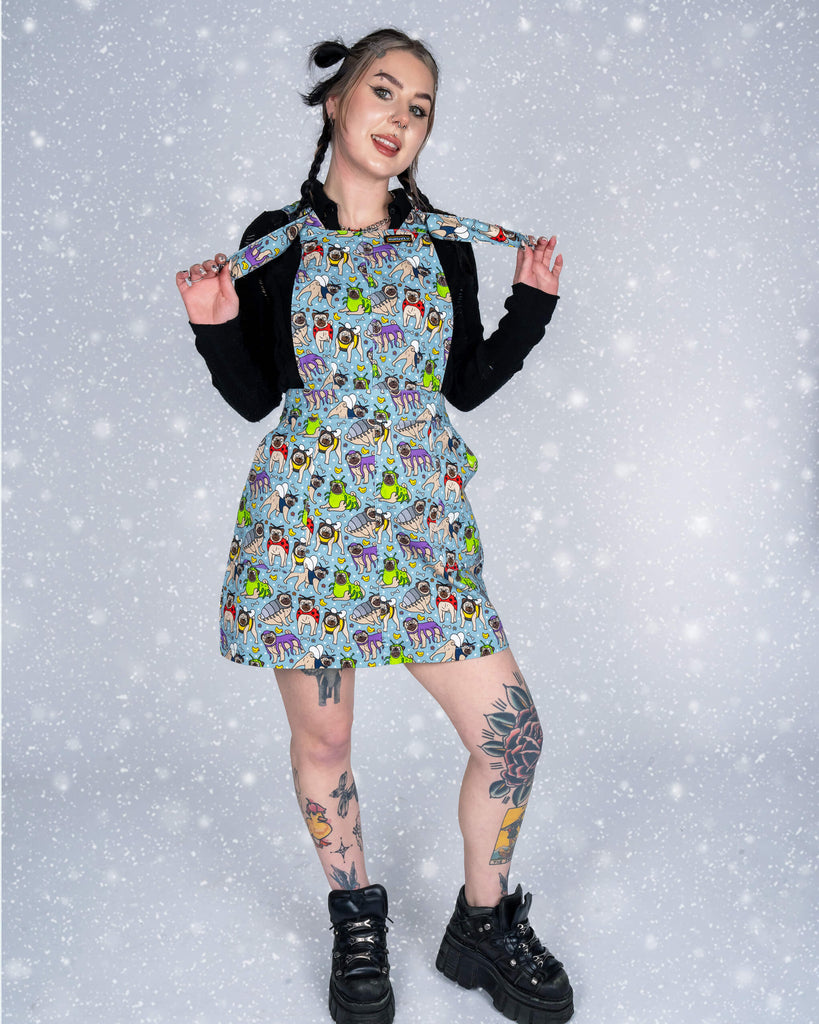 lo poses with a playful smile, holding the straps of the Pug Bugs pinafore dress. The vibrant design, with pugs in bug costumes, pops against her black top and the snow-effect backdrop.