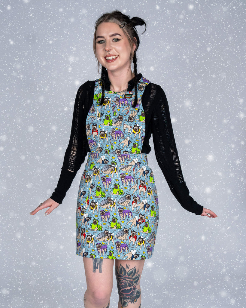 Flo is modelling a pinafore dress in the Pug Bugs print by Waffle Mama Design for Run & Fly. The dress features adorable pugs dressed as insects like ladybirds and bees. Flo pairs it with a black top and chunky black boots, standing against a snow-effect backdrop.