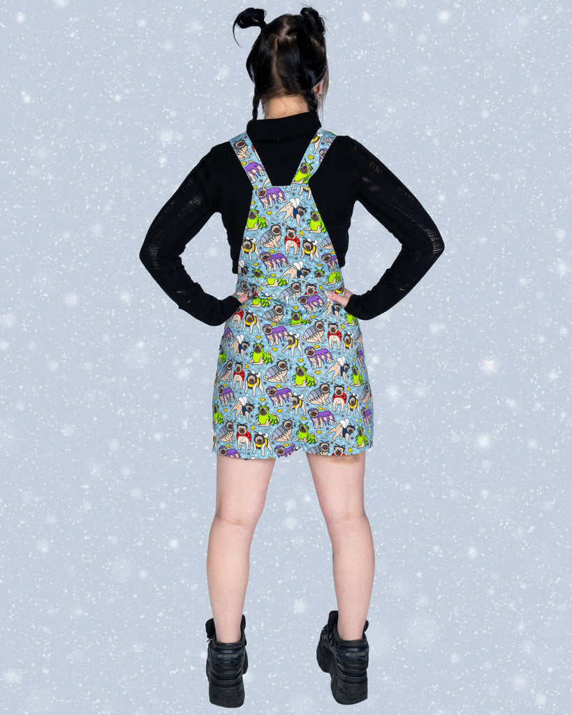 Flo shows off the back of the Pug Bugs pinafore dress, highlighting the adjustable straps and playful pug print. She stands with her hands on her hips, dressed in a black top and chunky black boots.