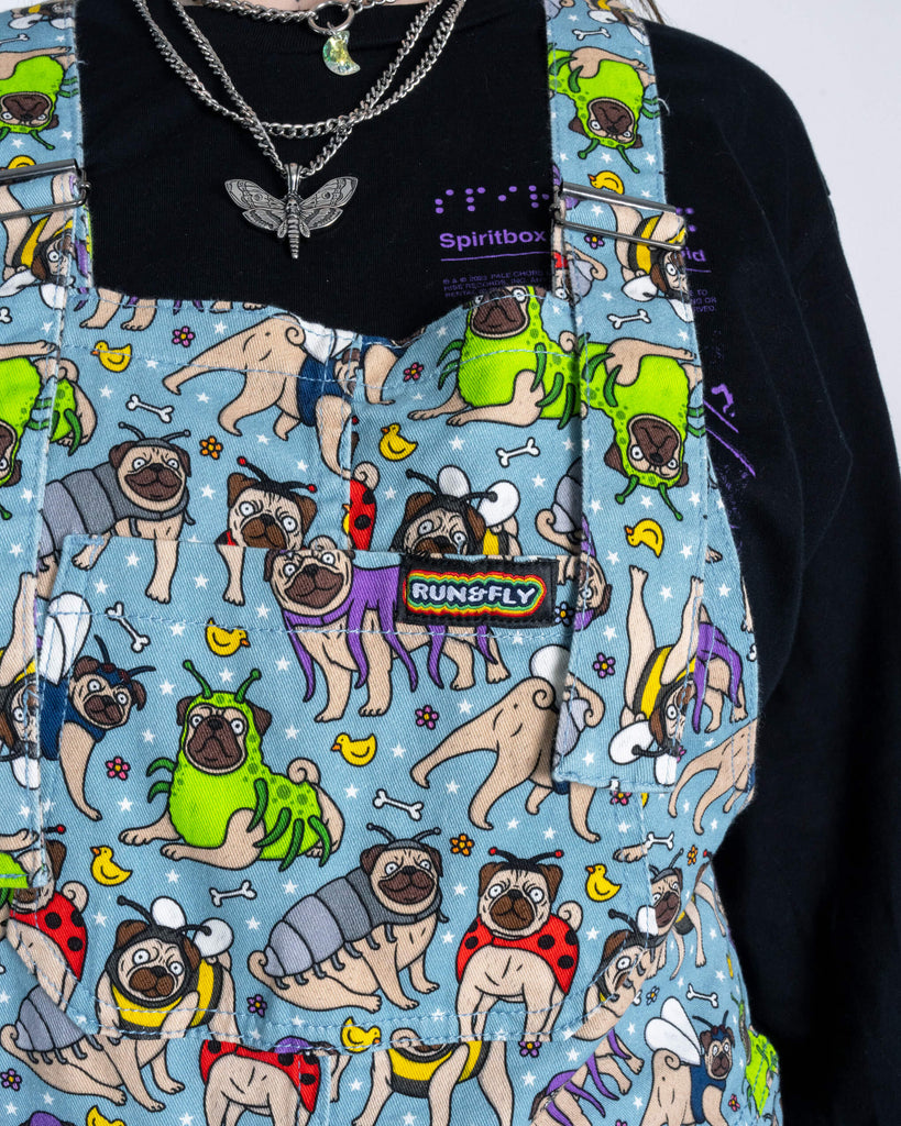 Close-up of Run & Fly ‘Pug Bugs’ dungarees, showing the detailed pug-and-bug print on a light blue background, with the Run & Fly rainbow label on the chest pocket.