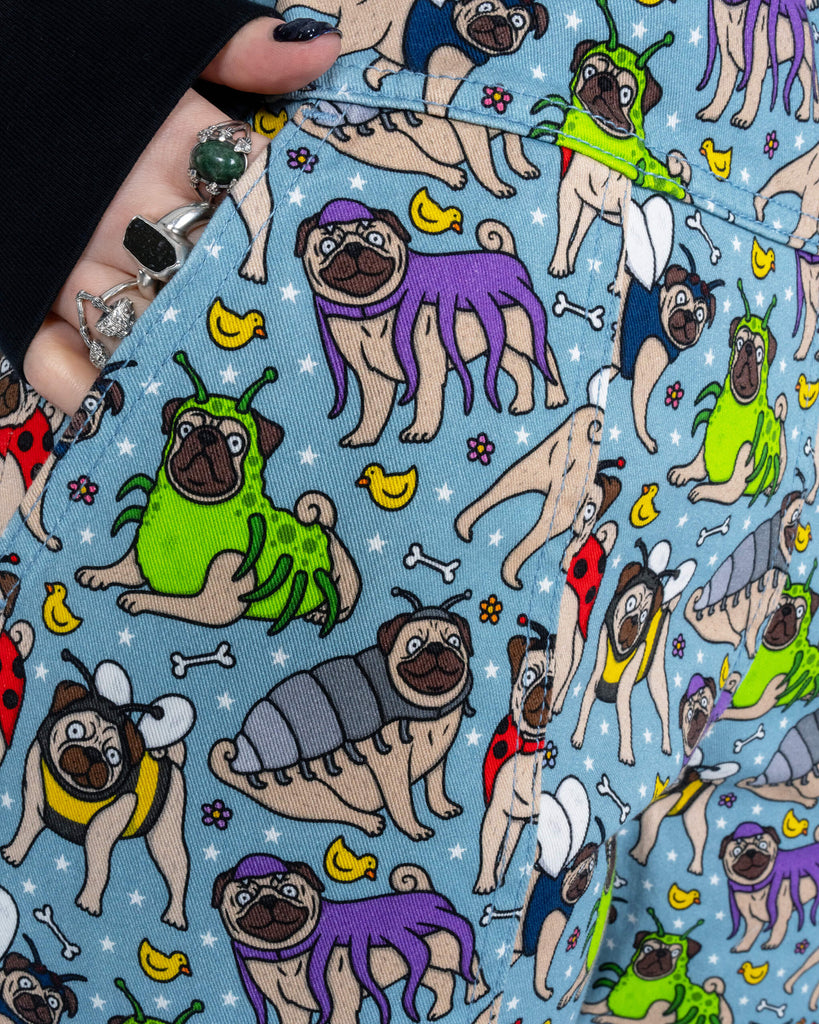 Close-up of Faeryn’s hand in the pocket of the Run & Fly ‘Pug Bugs’ dungarees, showcasing the playful print of pugs dressed as bugs with rubber ducks and stars in the background.