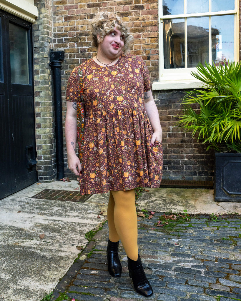 Full view of Maddie in the autumnal pumpkin print dress by Run & Fly, featuring a warm blend of oranges, browns, and greens designed by Jen James. The dress has pockets and falls just above the knees, paired with mustard tights and black ankle boots.