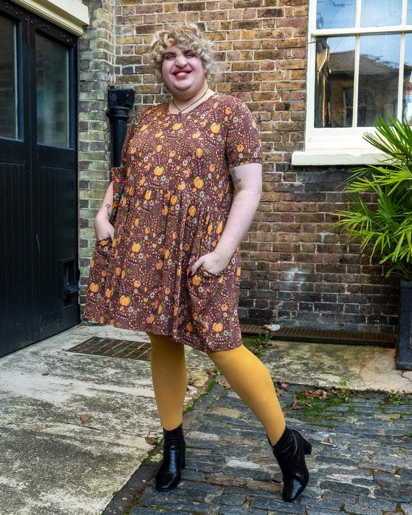 Maddie poses with a smile, showing off the flowing skirt of the pumpkin-patterned dress by Run & Fly, accented by mustard tights and black ankle boots for a seasonal look.