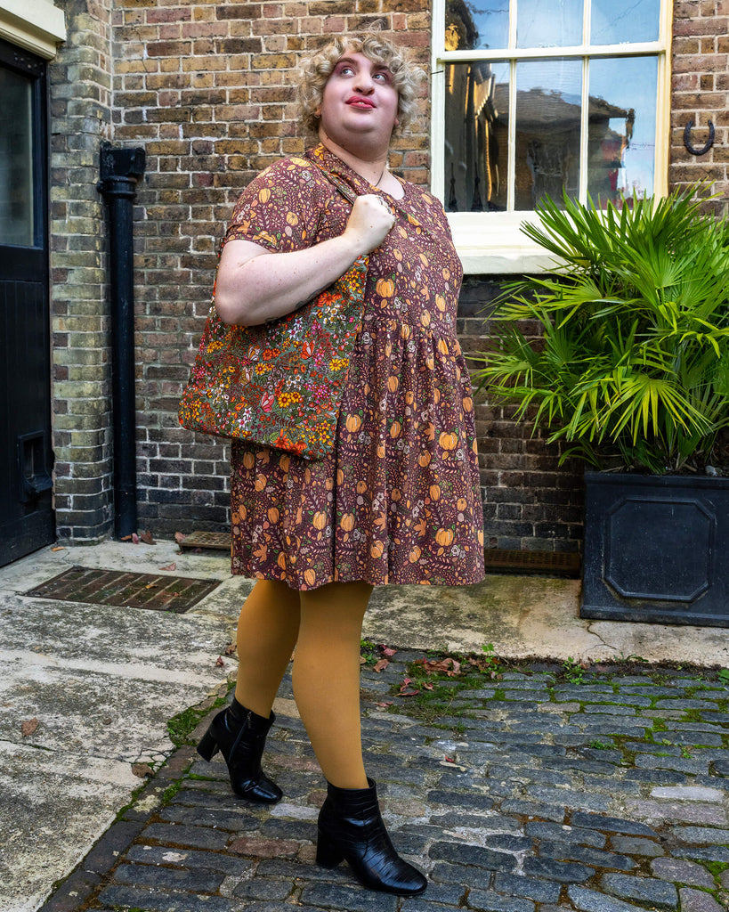 Maddie stands outdoors, wearing the Run & Fly pumpkin dress, holding a matching tote bag with a similar autumnal design. The dress has short sleeves and a relaxed fit, perfect for a cosy autumn look.