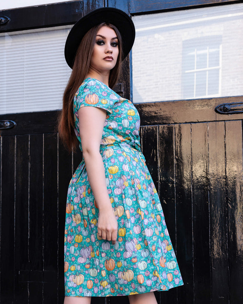 Charlotte is stood outside wearing the Halloween: Pumpkin Patch Stretch Belted Tea Dress with Pockets with a witchy styled black wide brim hat. She is facing away and looking over her shoulder to camera with both arms resting by her side. The pumpkin patch tea dress is an a bright turquoise base with pastel pink, purple, yellow, orange & mint pumpkins repeating all over with white sparkles. The dress has a round neck, is short sleeved, has a pleated skirt and a detachable belt tie.