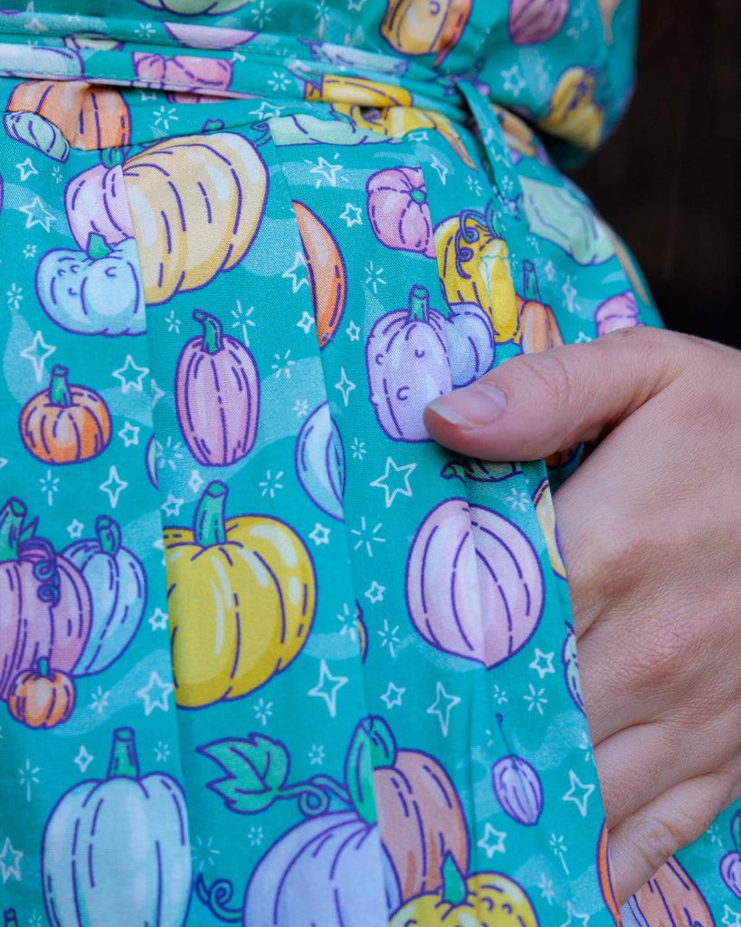 Close up of Charlotte's hand in the pocket of the Halloween: Pumpkin Patch Stretch Belted Tea Dress with Pockets. The pumpkin patch tea dress is an a bright turquoise base with pastel pink, purple, yellow, orange & mint pumpkins repeating all over with white sparkles. The dress has a round neck, is short sleeved, has a pleated skirt and a detachable belt tie.