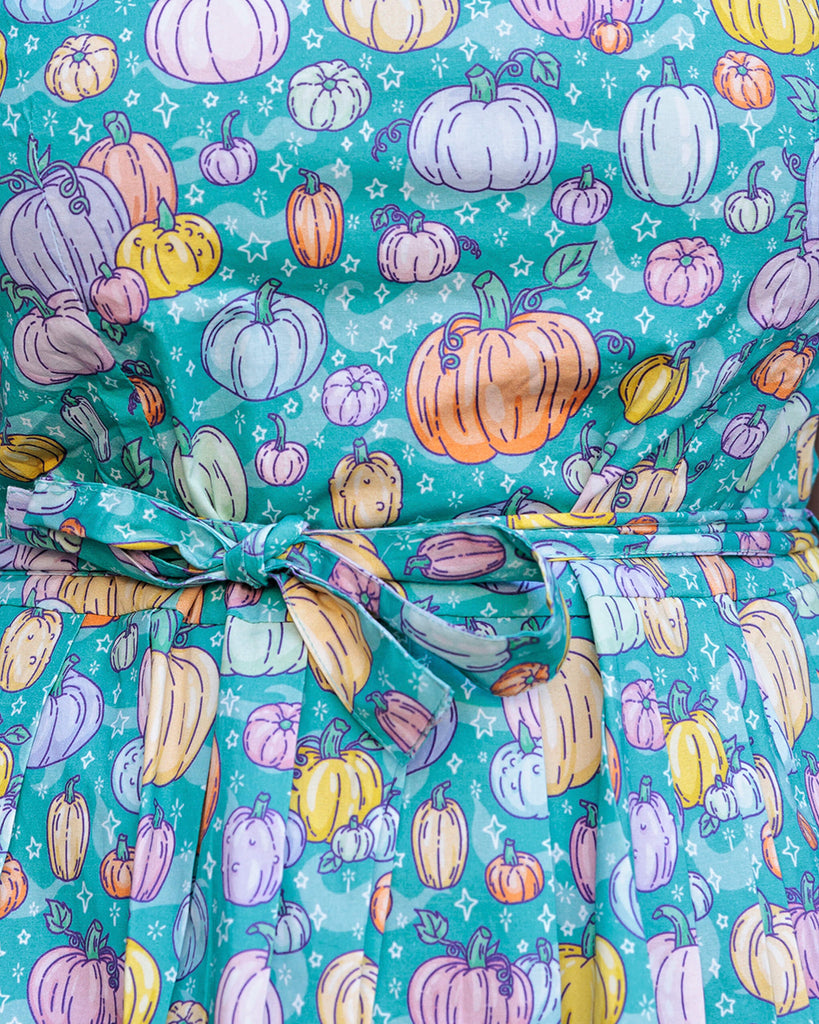 Close up of the Halloween: Pumpkin Patch Stretch Belted Tea Dress with Pockets. The pumpkin patch tea dress is an a bright turquoise base with pastel pink, purple, yellow, orange & mint pumpkins repeating all over with white sparkles. The dress has a round neck, is short sleeved, has a pleated skirt and a detachable belt tie.