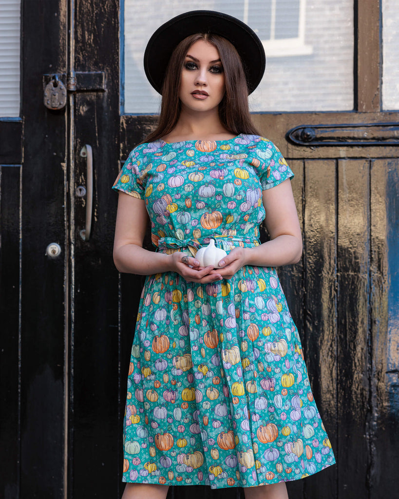 Charlotte is stood outside wearing the Halloween: Pumpkin Patch Stretch Belted Tea Dress with Pockets with a witchy styled black wide brim hat. She is facing forward holding a ceramic white pumpkin at her waist. The pumpkin patch stretch belted tea dress is an a bright teal turquoise base with pastel pink, purple, yellow, orange and mint pumpkins repeating all over with white sparkles. The dress has a round neck, is short sleeved, has a pleated skirt and a detachable belt tie.