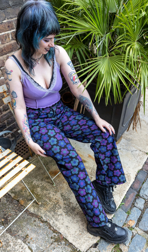 Faeryn a femme non binary person with blue and black hair and dark make up is wearing floral jeans and lilac top in a Hove mews they are sat down on a white chair