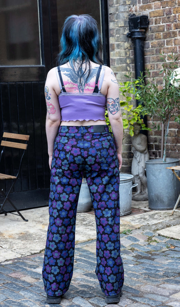 Faeryn a femme non binary person with blue and black hair and dark make up is wearing floral jeans and lilac top in a Hove mews they are facing the back to show off the doors