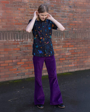 Jack is stood in front of a brick wall wearing the purple corduroy bell bottom flares with the cosmic space shirt and black boots. They are posing with their hands in their hair and are looking down.