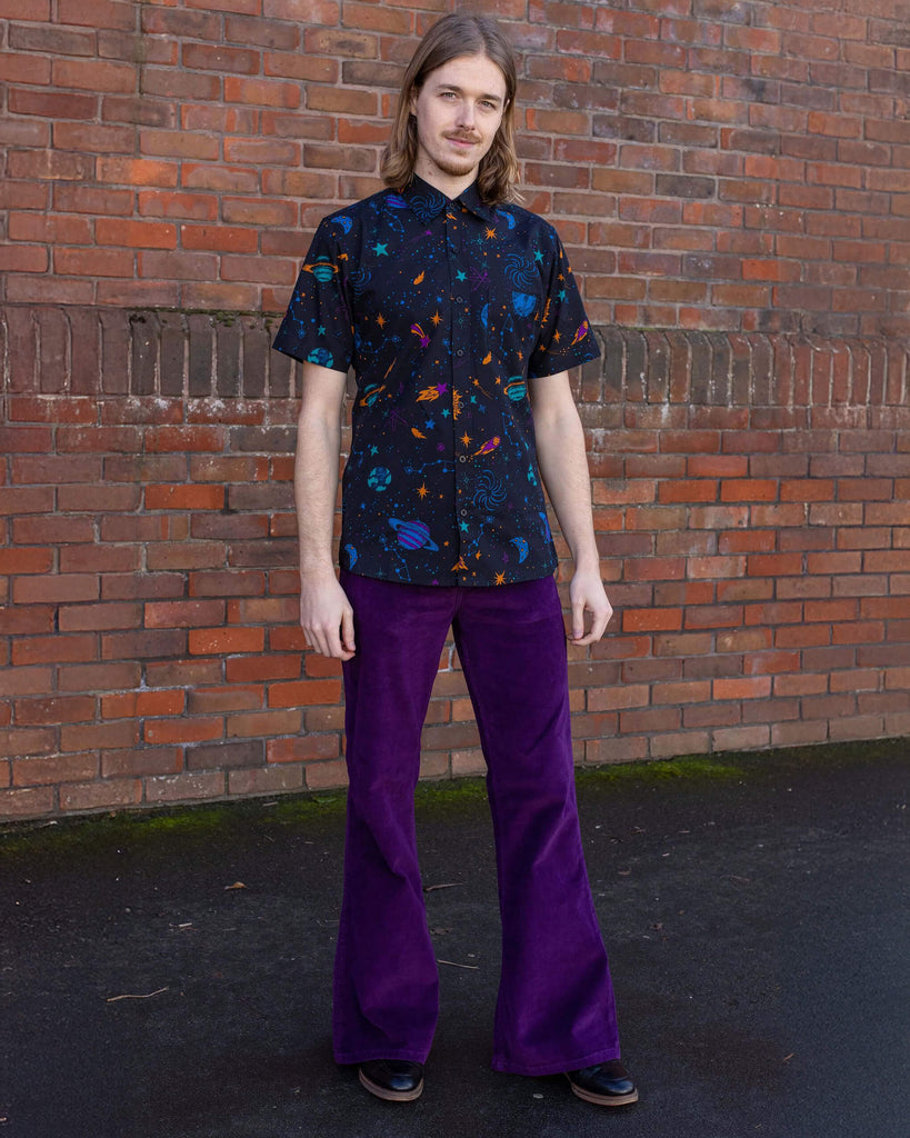 Jack is stood in front of a brick wall wearing the purple corduroy bell bottom flares with the cosmic space shirt and black boots. They are facing forward posing with both arms resting by their side and smiling to camera.