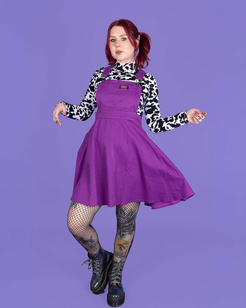 model with red hair wearing a purple flared pinafore paired with a long sleeve cow print turtle neck, fishnet tights and boots. Model is posing while swishing the skirt of the pinafore in front of a purple background.