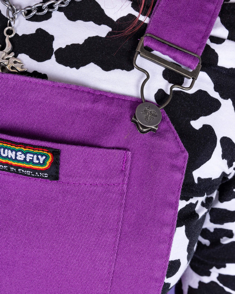 close up of model wearing a purple flared pinafore paired with a long sleeve cow print turtle neck. Close up shows the metal buckles and Run & Fly logo on the front pocket of the pinafore.