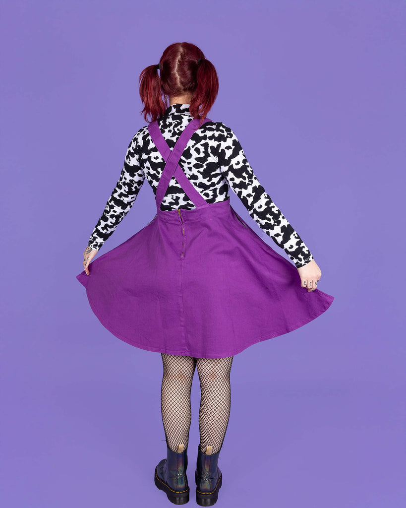 model with red hair wearing a purple flared pinafore paired with a long sleeve cow print turtle neck, fishnet tights and boots. Model is standing with their back to the camera and holding the skirt of the pinafore with both hands. The background of the photo is purple.