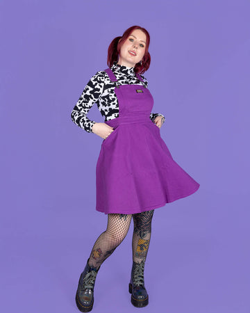 model with red hair wearing a purple flared pinafore paired with a long sleeve cow print turtle neck, fishnet tights and boots. Model is smiling with her hands in the pinafore pockets in front of a purple background.