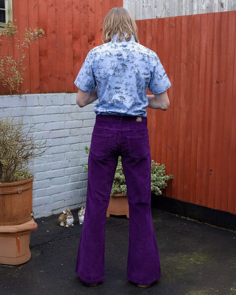 Jack is stood in front of a panelled brick and wood wall wearing the purple corduroy bell bottom flares with the blue safari shirt and black boots. They are facing away from the camera posing with both hands in front of them doing up the shirt buttons.