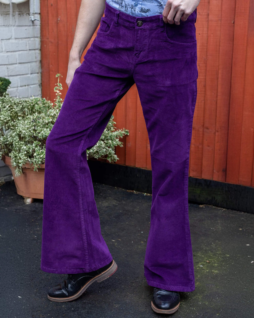  Jack is stood in front of a panelled brick and wood wall wearing the purple corduroy bell bottom flares with the blue safari shirt and black boots. They are facing forward posing with one hand pulling up the waist band and their leg bent to the side to highlight the wide flares. Photo is cropped from the waist down.