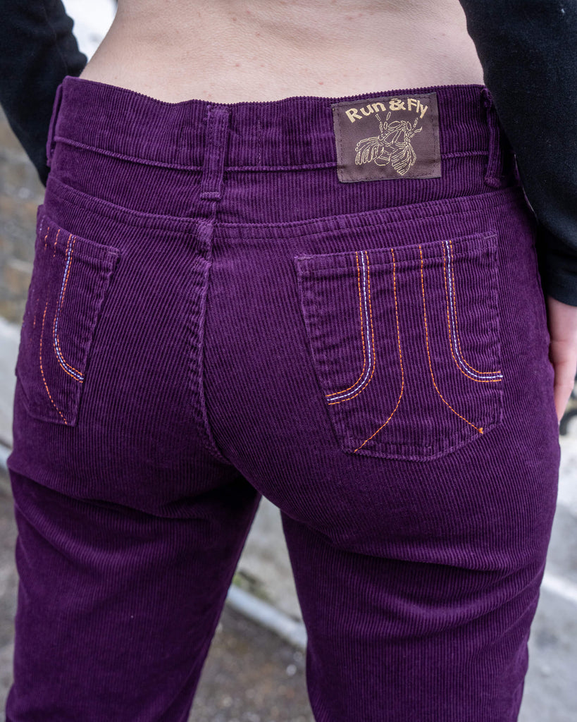 Back close-up of the Run & Fly Midnight Purple Corduroy Flares, featuring contrast orange stitching, back pockets, and a brown woven label with a gold bee design.