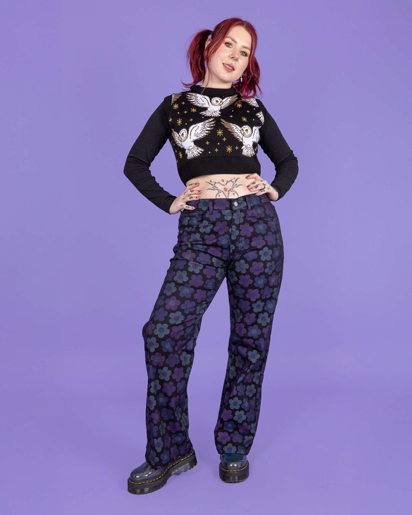 Florence a tattooed model with red hair is wearing DARK WASH 90's Black Magic Flower Wide Leg Stretch Jeans paired with a long sleeve black top, What a Hoot tank top and chunky boots. The black jeans have muted blue and purple flowers all over. The model is smiling at the camera with her hands on her hips and one leg turned outwards. The background of the photo is purple.