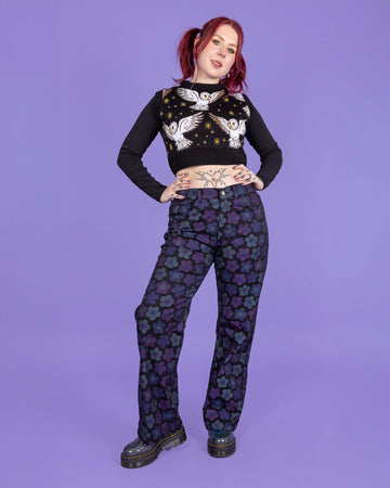 Florence a tattooed model with red hair is wearing DARK WASH 90's Black Magic Flower Wide Leg Stretch Jeans paired with a long sleeve black top, What a Hoot tank top and chunky boots. The black jeans have muted blue and purple flowers all over. The model is smiling at the camera with her hands on her hips and one leg turned outwards. The background of the photo is purple.