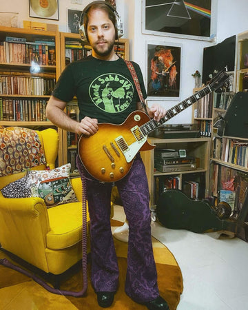 Stefano is stood in their 70s themed lounge next to a yellow chair wearing the purple grape Hendrix paisley corduroy retro bell bottom flares with a black sabbath tee and black boots. They have a brown electric guitar on them with studio headphones on. They are facing forward posing with their hands resting on their guitar going across them whilst looking to camera.