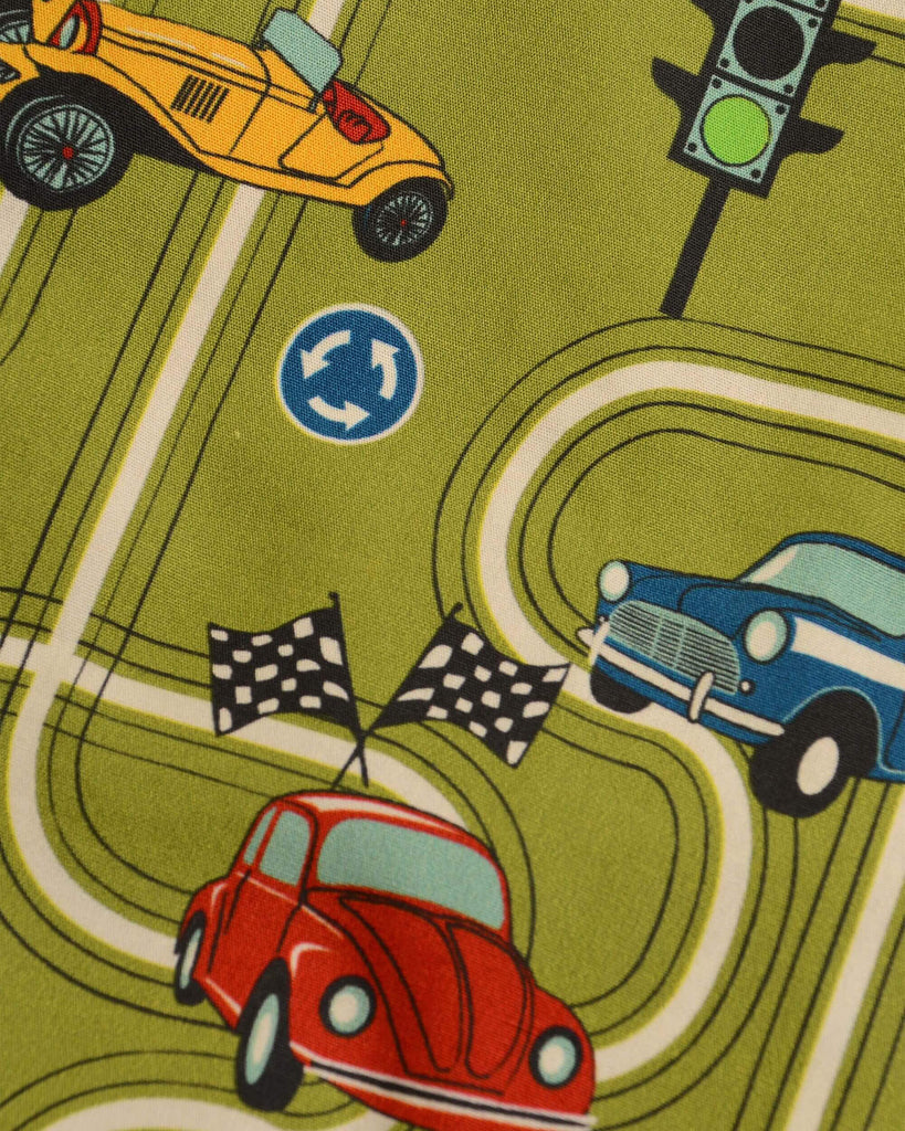 Close up shot of the race track car play button down long sleeve shirt fabric which showcases blue, yellow and red cars with a winding track, car signs and traffic lights on a kiwi green base colour.