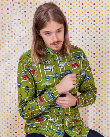 Jack is sat on a yellow stool in front of a multicoloured polka dot background wearing the race track car play button down long sleeve shirt with denim jeans. They are facing forward and posing doing up the sleeve cuff buttons whilst looking down to the right. Photo is cropped from the hips up.