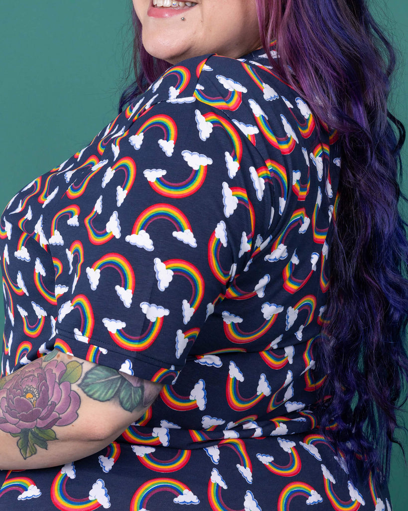 The Over the Rainbow & Clouds Short Sleeve T Shirt worn by a femme alternative tattooed model with rainbow glasses, long pink to purple hair and the matching cycle shorts on a teal studio background. She is facing left smiling with her arms resting on her hips. The navy blue base tshirt has repeating rainbow arches with white clouds.