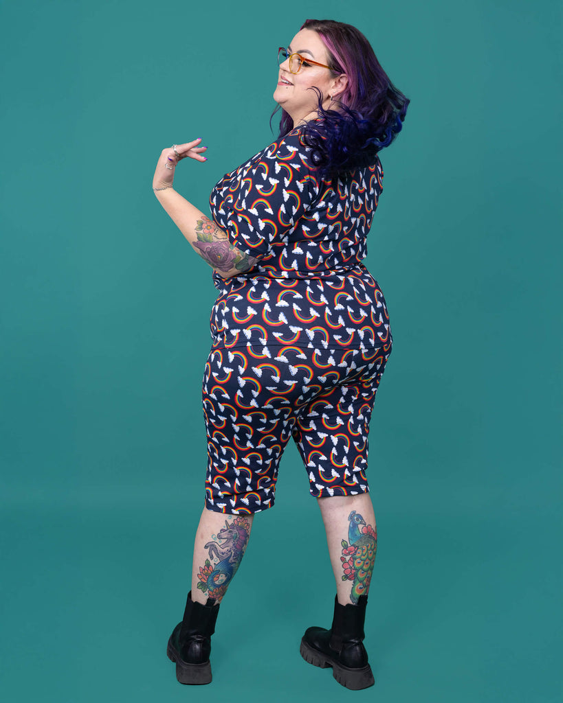The Over the Rainbow & Clouds Stretch Cycle Shorts worn by a femme alternative tattooed model with rainbow glasses, long pink to purple hair, the matching tshirt and black boots on a teal studio background. She is facing away flicking her hair over her shoulder and smiling to the left. The navy blue base shorts has repeating rainbow arches with white clouds.