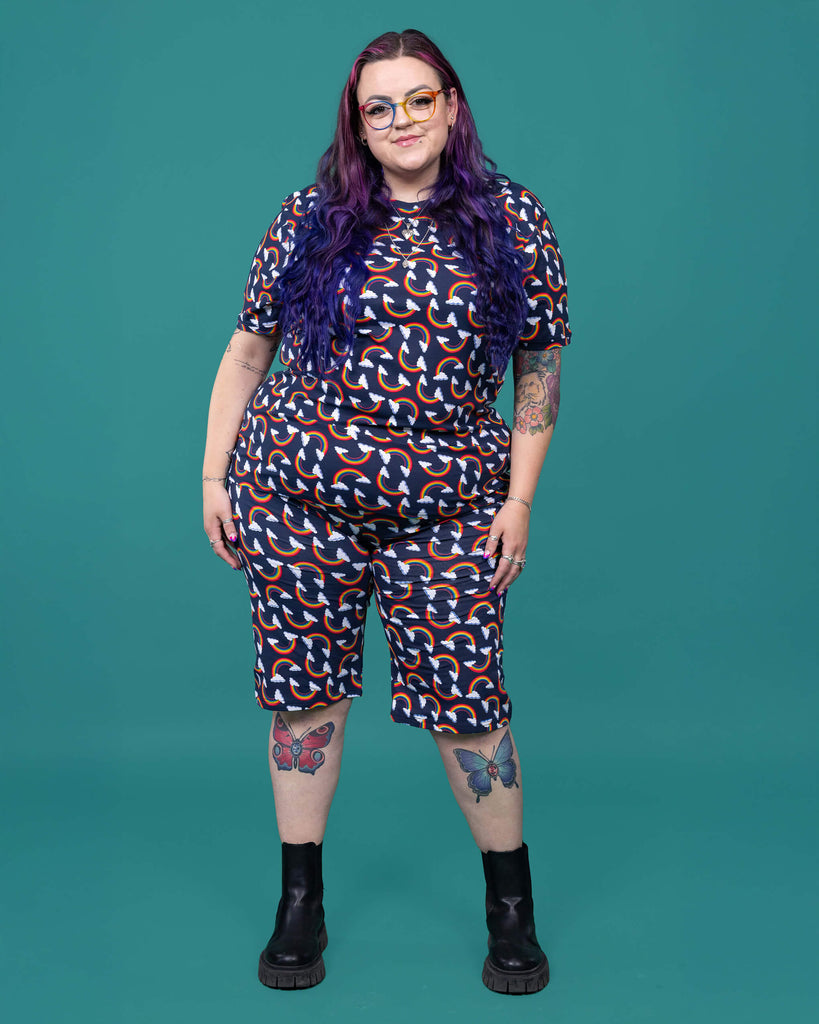 The Over the Rainbow & Clouds Stretch Cycle Shorts worn by a femme alternative tattooed model with rainbow glasses, long pink to purple hair, the matching tshirt and black boots on a teal studio background. She is facing forward smiling with both arms resting by her side. The navy blue base shorts has repeating rainbow arches with white clouds.