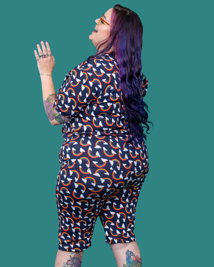 The Over the Rainbow & Clouds Short Sleeve T Shirt worn by a femme alternative tattooed model with rainbow glasses, long pink to purple hair and the matching cycle shorts on a teal studio background. She is facing away from camera laughing looking up over her left shoulder with her arm up. The navy blue base tshirt has repeating rainbow arches with white clouds.