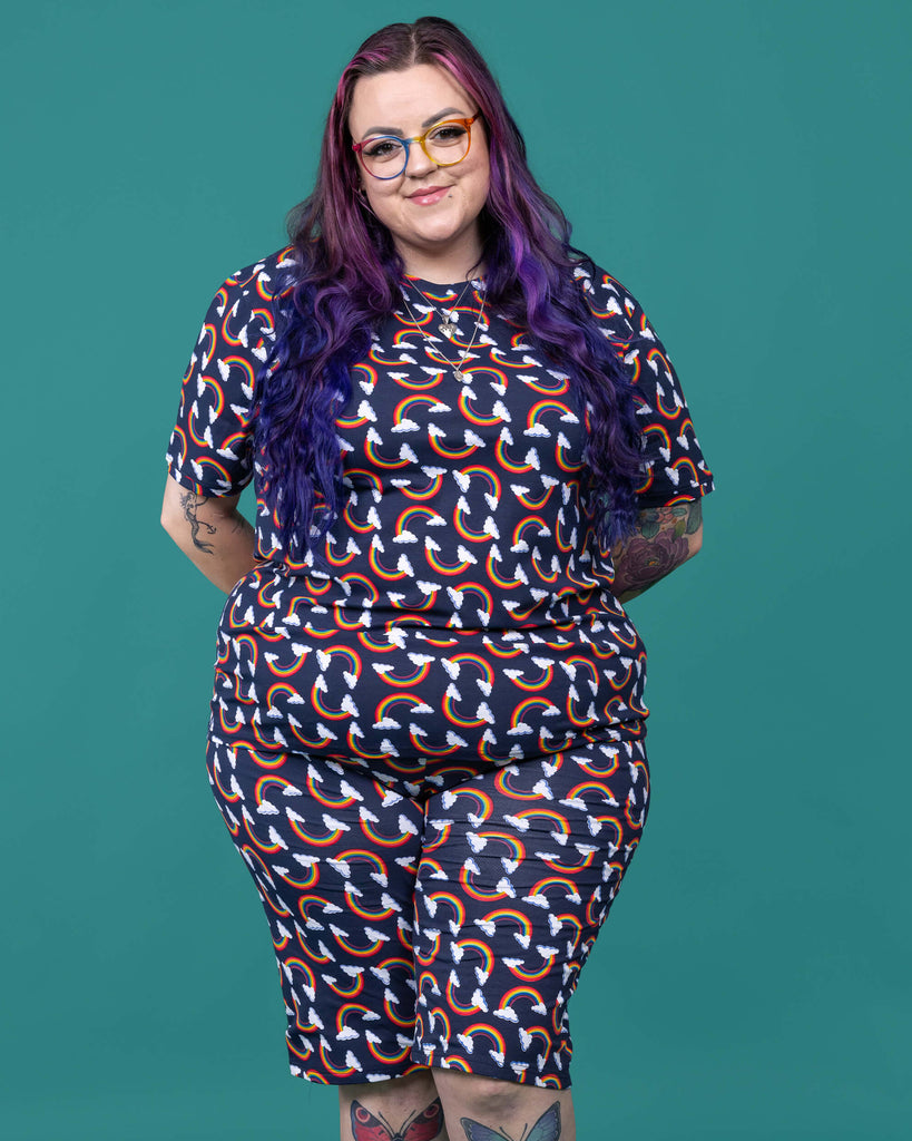 The Over the Rainbow & Clouds Stretch Cycle Shorts worn by a femme alternative tattooed model with rainbow glasses, long pink to purple hair, the matching tshirt and black boots on a teal studio background. She is facing forward smiling with both hands resting behind her back. The navy blue base shorts has repeating rainbow arches with white clouds.