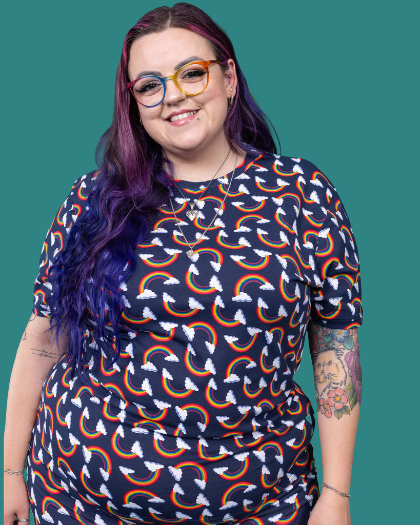 The Over the Rainbow & Clouds Short Sleeve T Shirt worn by a femme alternative tattooed model with rainbow glasses, long pink to purple hair and the matching cycle shorts on a teal studio background. She is facing forward smiling with both hands resting by her sides. The navy blue base tshirt has repeating rainbow arches with white clouds.