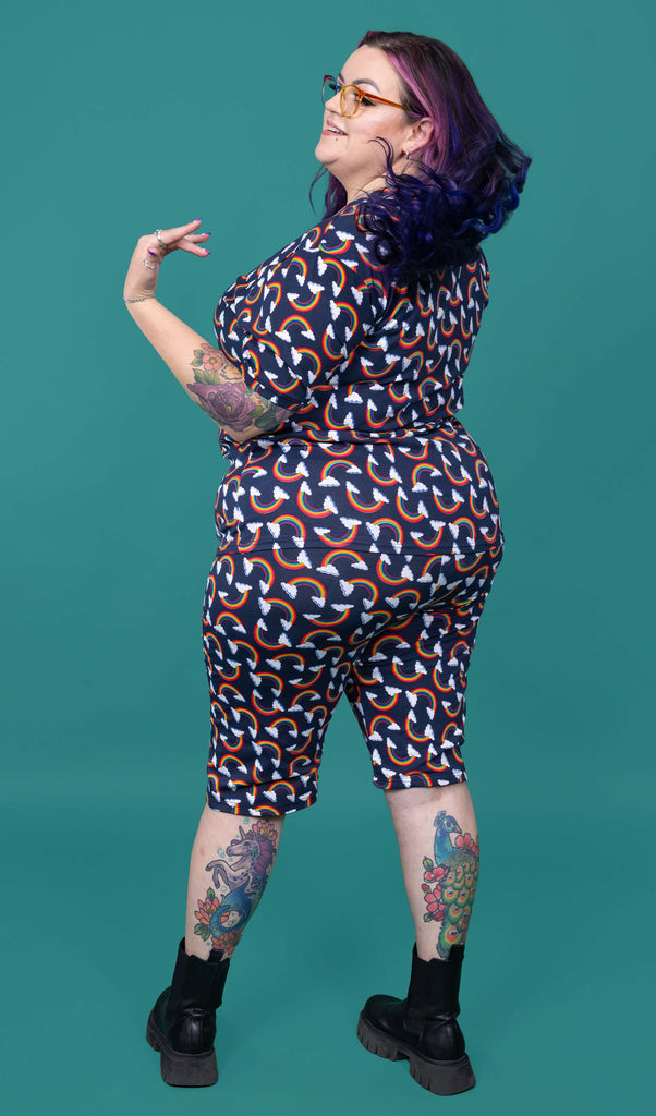 The Over the Rainbow & Clouds Stretch Cycle Shorts worn by a femme alternative tattooed model with rainbow glasses, long pink to purple hair, the matching tshirt and black boots on a teal studio background. She is facing away flicking her hair over her shoulder and smiling to the left. The navy blue base shorts has repeating rainbow arches with white clouds.