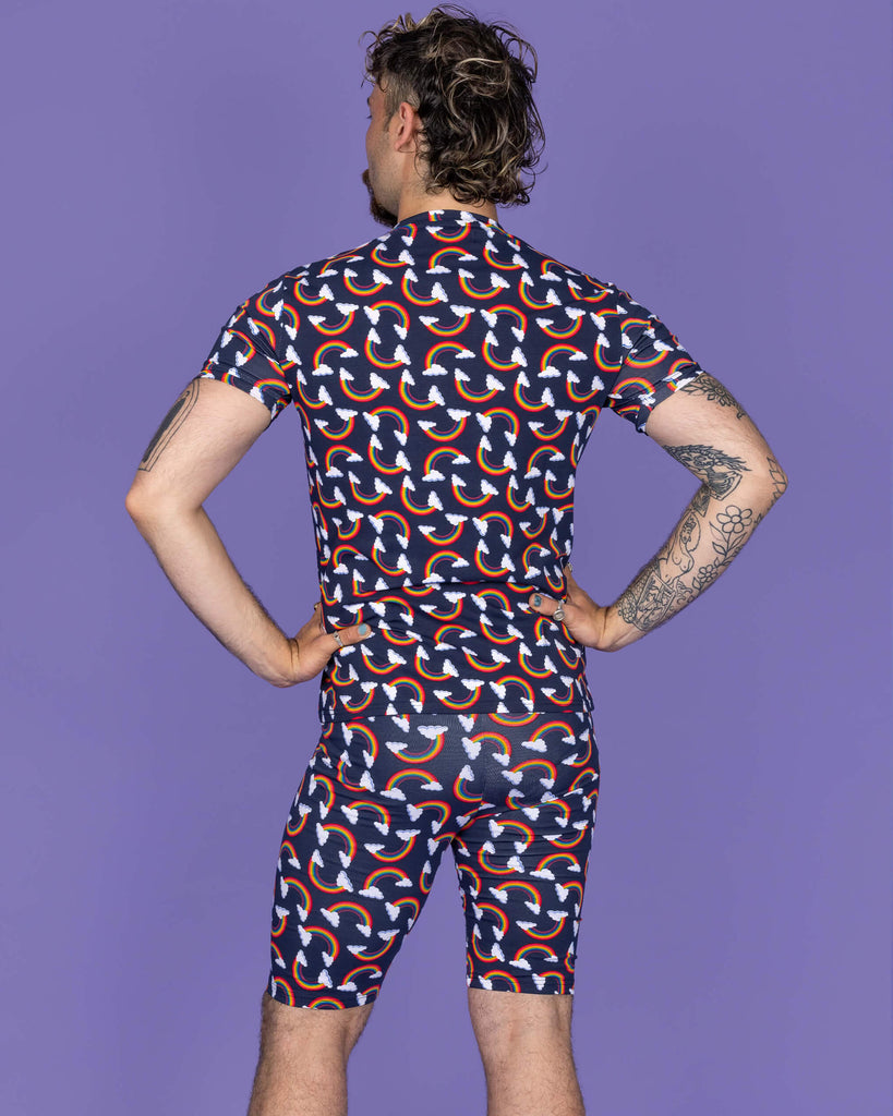 The Over the Rainbow & Clouds Stretch Cycle Shorts worn by a masc non binary person with a brown curly mullet, the matching top and white trainers on a purple studio background. They are facing away with both hands resting on their hips. The navy blue base shorts has repeating rainbow arches with white clouds.