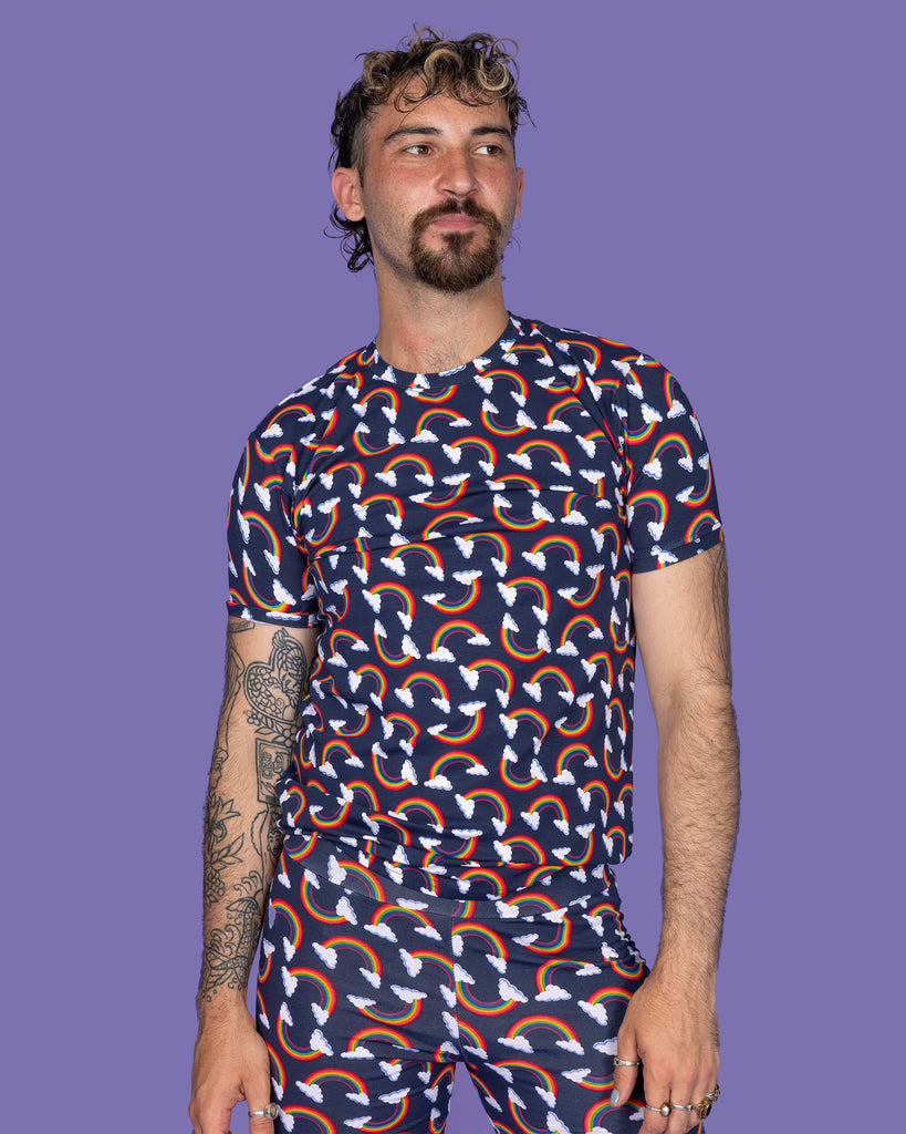 The Over the Rainbow & Clouds Short Sleeve T Shirt worn by a masc non binary person with a brown curly mullet and the matching cycle shorts on a purple studio background. They are facing forward smiling looking off to the right with both hands resting by their sides. The navy blue base tshirt has repeating rainbow arches with white clouds.