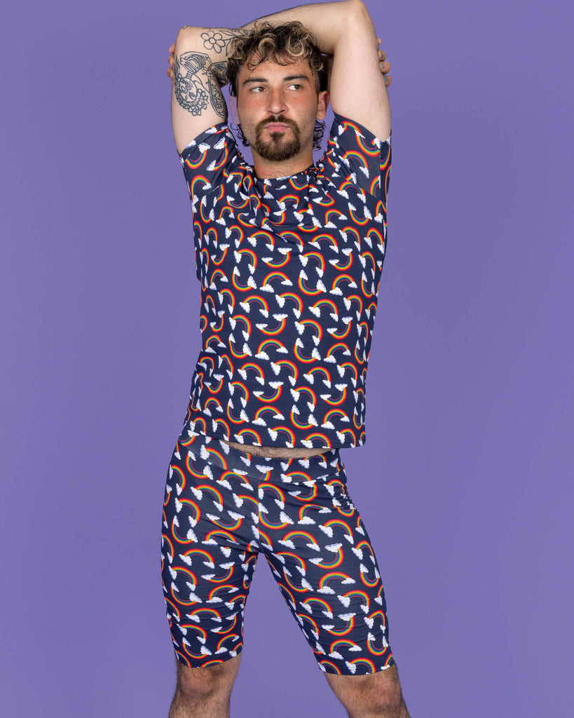 The Over the Rainbow & Clouds Stretch Cycle Shorts worn by a masc non binary person with a brown curly mullet, the matching top and white trainers on a purple studio background. They are facing forward pouting with both arms folded above their head. The navy blue base shorts has repeating rainbow arches with white clouds.