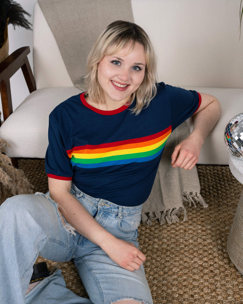 The model sits cross-legged, wearing the Navy Ringer Retro Rainbow Striped T Shirt by Run & Fly. The bold rainbow stripe and red ringer details give it a cheerful, retro feel. Styled with ripped light-wash jeans, this ethical, slow fashion piece is shown in a warm, relaxed setting.